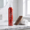 Red foam hair product bottle with a wooden comb and white towel on a marble surface.